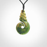 This picture shows a hook shaped pendant, hand carved from New Zealand flower jade. The top of the hook, the shaft, is a twist shape. We will provide this with an adjustable plaited cord. The cord is a adjustable and is a black colour. Twist and koru designs are also displayed in this stunning New Zealand Marsden Flower Jade pendant. Hand made by Ana Krakosky.